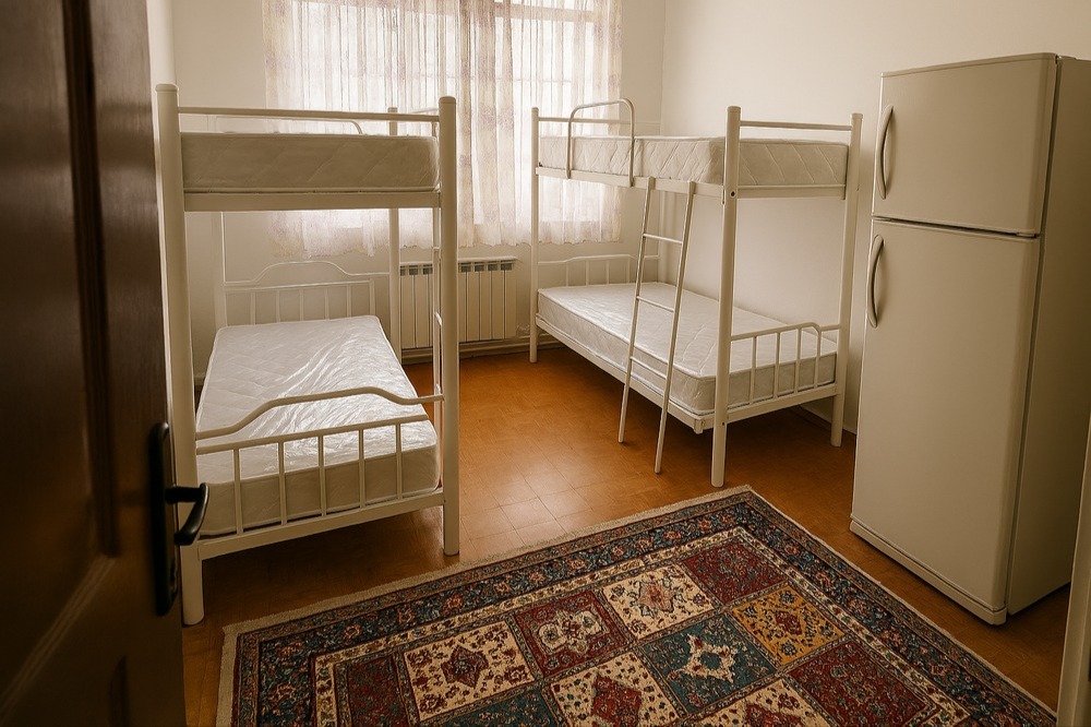 اتاق خواب مینیمال با تخت دوطبقه فلزی سفید، یخچال کوچک و پنجره نورگیر با پرده حریر گلدار خوابگاه دخترانه در کرج اتاق خواب مدرن و مینیمال با دو تخت دوطبقه فلزی سفید، تشکهای نو با پوشش پلاستیکی، یخچال سفید کوچک در گوشه اتاق، پنجره بزرگ با پرده حریر گلدار، کفپوش پارکت چوبی و فرش سنتی رنگارنگ؛ مناسب برای اقامت کوتاهمدت و خوابگاه